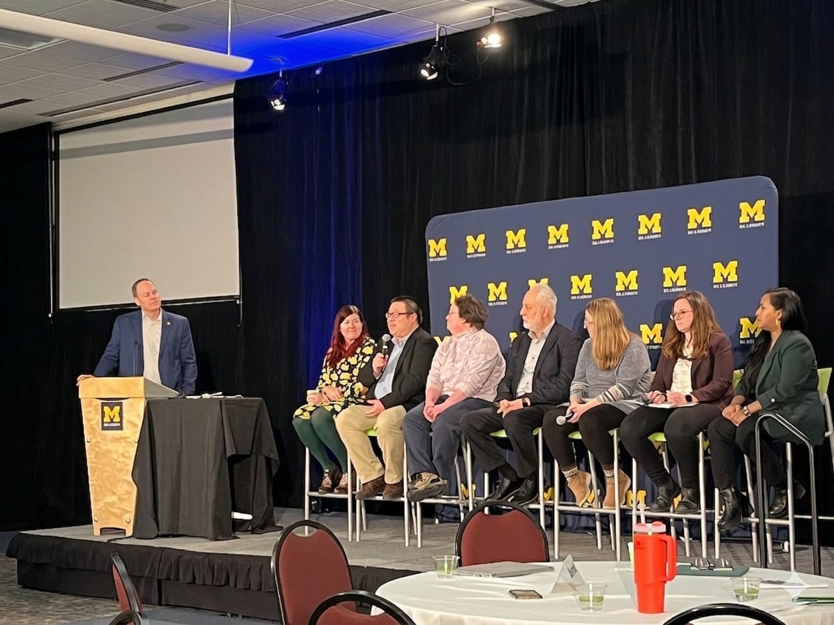 A University of Michigan panel discussion with one speaker at a podium and seven others seated on stage.