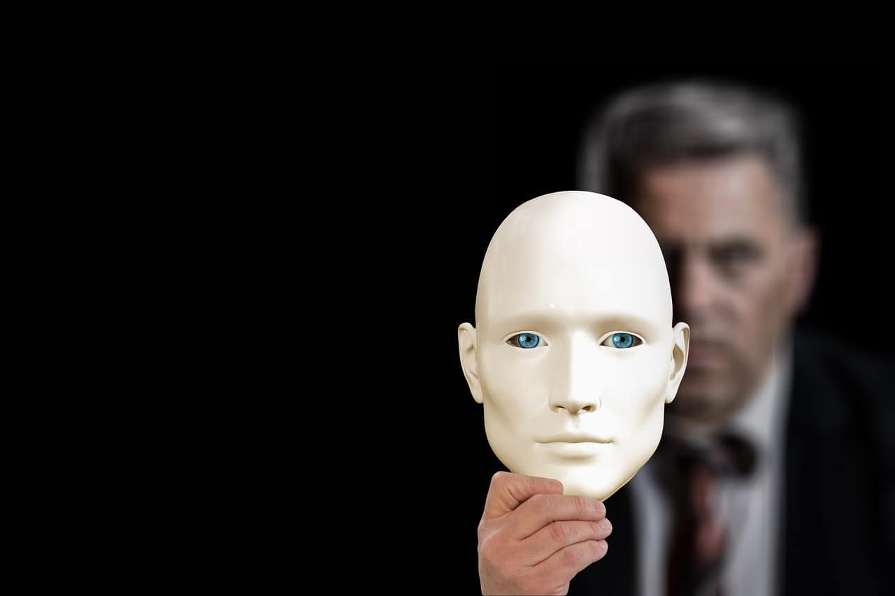 A man in a suit holds up a smooth, expressionless white mask with blue eyes, obscuring their own face. The actual person's face is blurred in the background against a black backdrop, creating a visual contrast between human identity and artificial or anonymous representation.