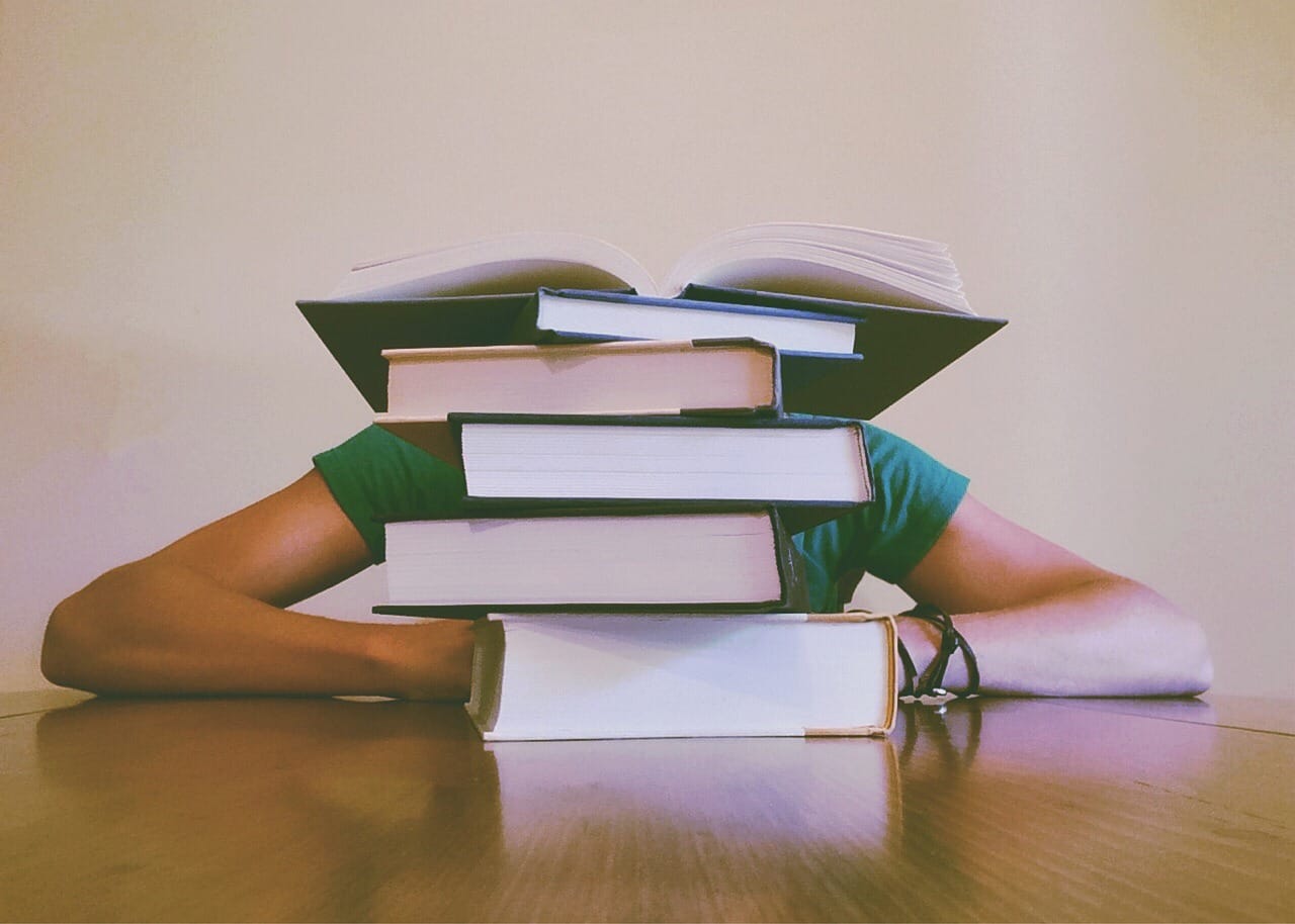 Person slumped at a table behind a tall stack of books.