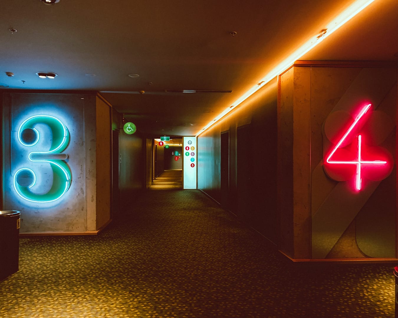 Dimly lit movie theater hallway with a large neon number “3” in blue on the left wall and “4” in red on the right, indicating theater numbers; additional signage and glowing icons are visible deeper down the corridor.