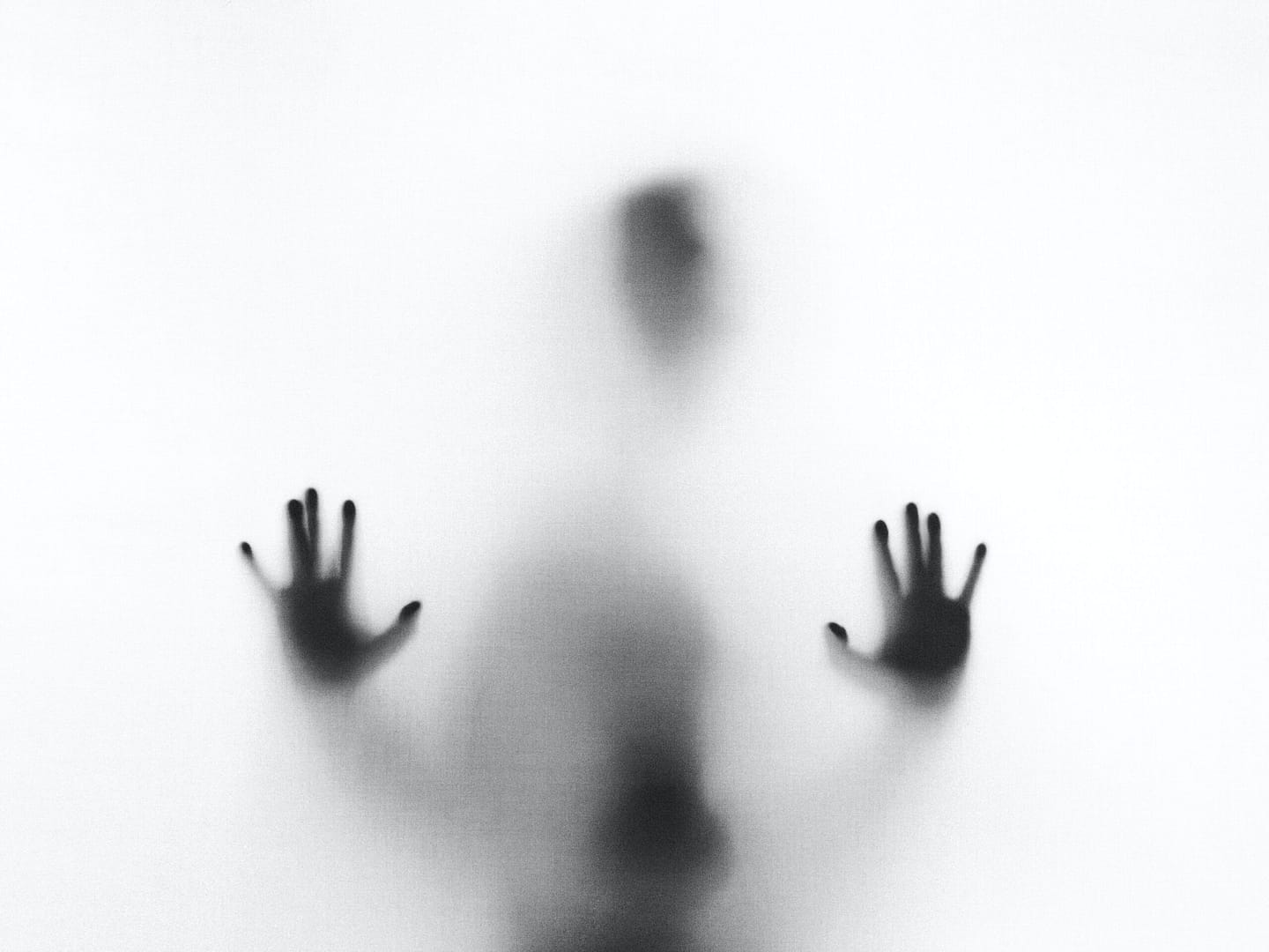 A black and white photo of a blurred human figure behind a translucent surface, with both hands pressed against it. The figure’s face and body are indistinct, creating an eerie or mysterious atmosphere.