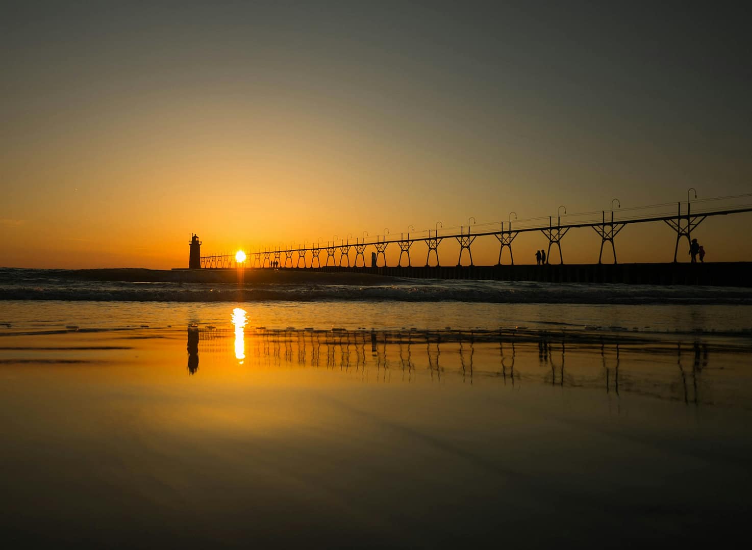 view of the horizon with a pedestrian foot path to a lighthouse in the distance, with the sun low in the sky reflecting on the water.