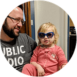 Circular crop of a bearded man holding a blond haired child who is wearing blue framed sunglasses