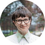 Circular crop of a portrait of a woman with short hair wearing black rimed glasses against an outdoor background