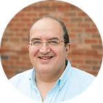 Circular crop of a portrait of a man wearing a blue shirt and glasses against a brick background