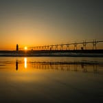 view of the horizon with a pedestrian foot path to a lighthouse in the distance, with the sun low in the sky reflecting on the water.