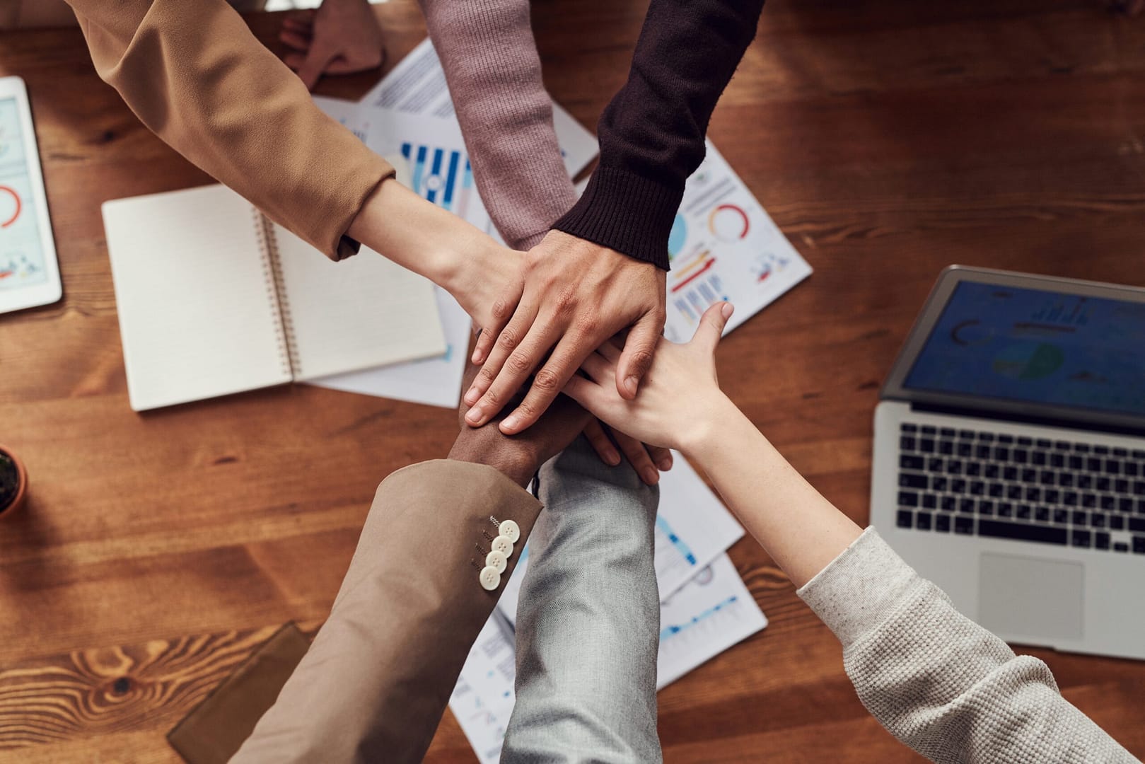 Top-down view of six hands stacked together in a gesture of teamwork and unity, surrounded by laptops, notebooks, and charts on a wooden table, suggesting a collaborative office or project setting.