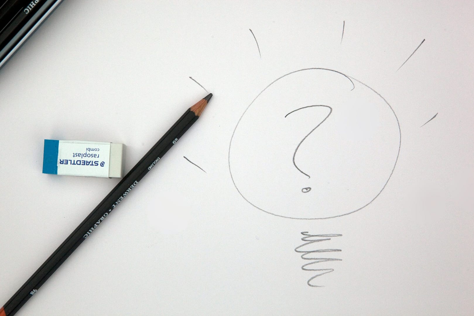 A close-up image of a pencil and a Staedtler eraser placed on a white sheet of paper. The paper features a hand-drawn light bulb shape with a question mark inside, symbolizing curiosity or the process of generating ideas. Radiating lines and a squiggly filament reinforce the light bulb concept, suggesting a moment of creative thinking or inquiry.