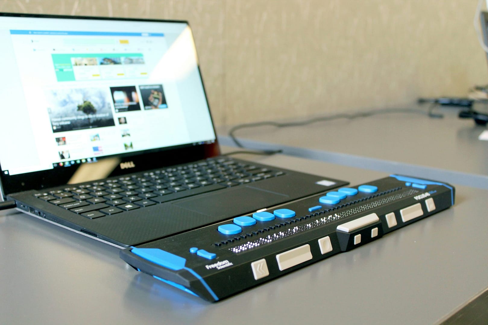 A laptop displaying a website sits on a table next to a refreshable braille display with blue and white tactile controls.