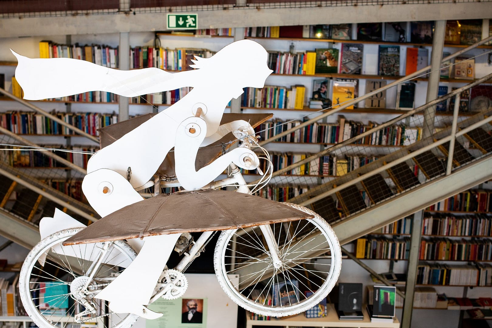 White sculpture of a person with flowing hair and scarf riding a whimsical flying bicycle, suspended in a bookstore with bookshelves and staircases in the background—blending imagination and knowledge in a literary space.