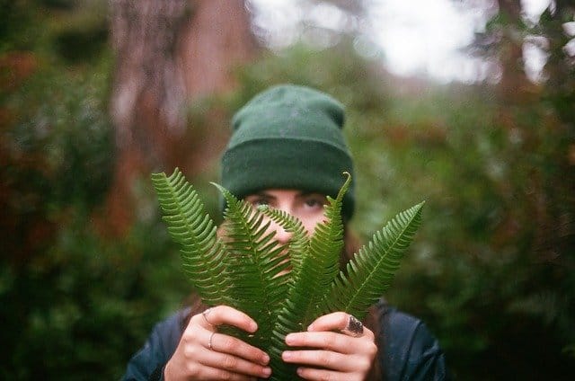 A woman peers from behind green leaves that she holds in her hands. Evokes a sense of hiding.