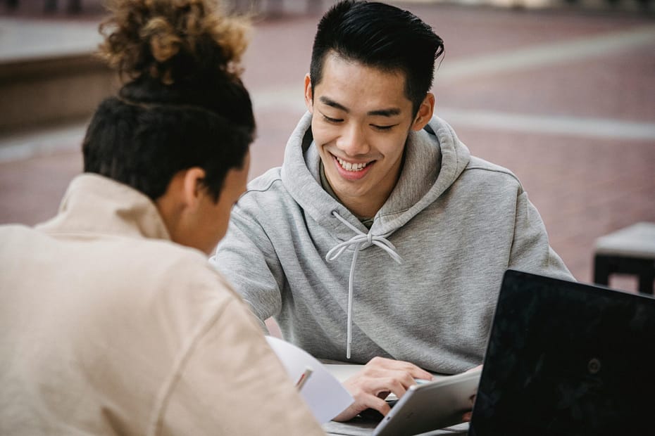 Two students sit together at an outdoor table, smiling and working collaboratively. One wears a gray hoodie and looks at a tablet, while the other, whose back is mostly to the camera, wears a beige jacket and appears to be taking notes on paper. A laptop is open on the table in front of them. The setting is relaxed and conducive to learning or studying.