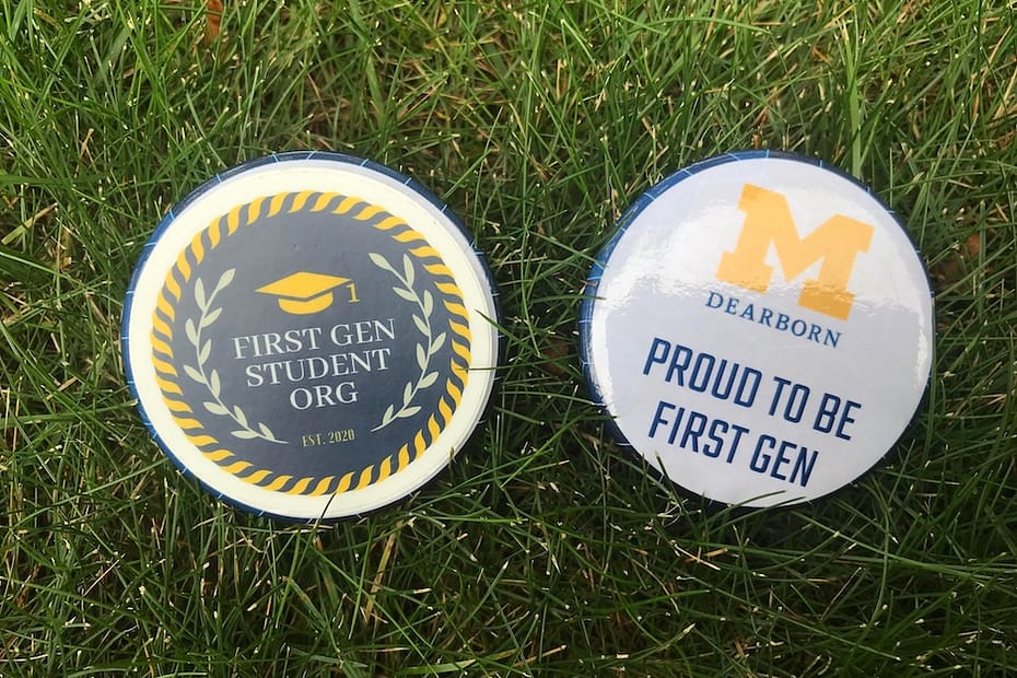 Two circular buttons lying on green grass. The button on the left features a dark blue background with a yellow graduation cap, laurel wreath design, and the text "FIRST GEN STUDENT ORG" and "EST. 2020." The button on the right has a white background with the University of Michigan-Dearborn's yellow block "M" logo and the text "PROUD TO BE FIRST GEN" in blue.