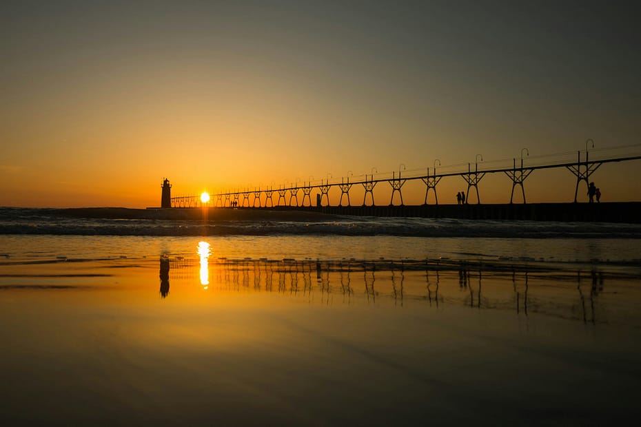 view of the horizon with a pedestrian foot path to a lighthouse in the distance, with the sun low in the sky reflecting on the water.