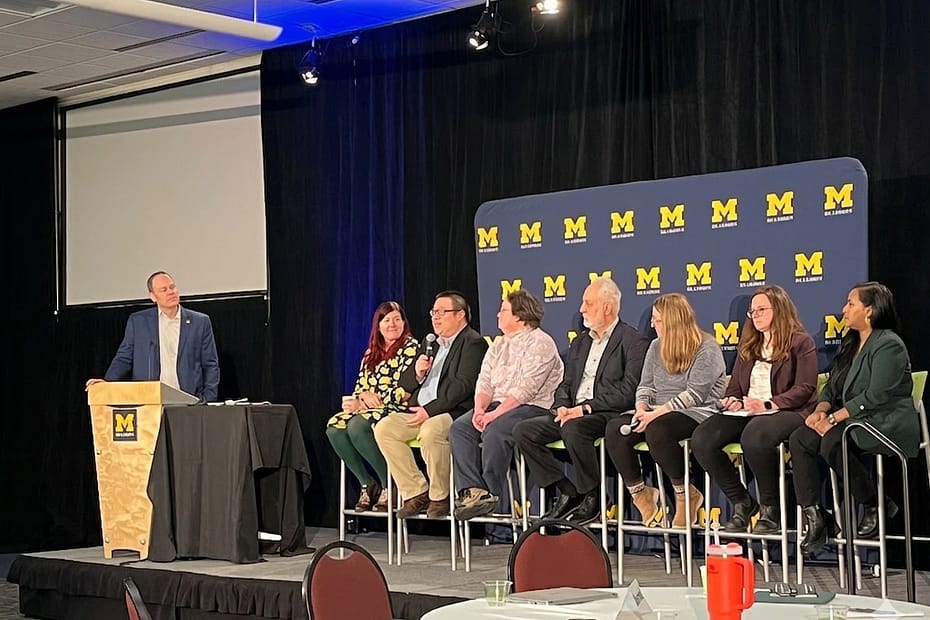A University of Michigan panel discussion with one speaker at a podium and seven others seated on stage.